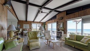 Un salón amplio con un gran ventanal con vistas al mar, vigas de madera en el techo y una zona de estar acogedora con un sofá y sillones de color verde con estampado.