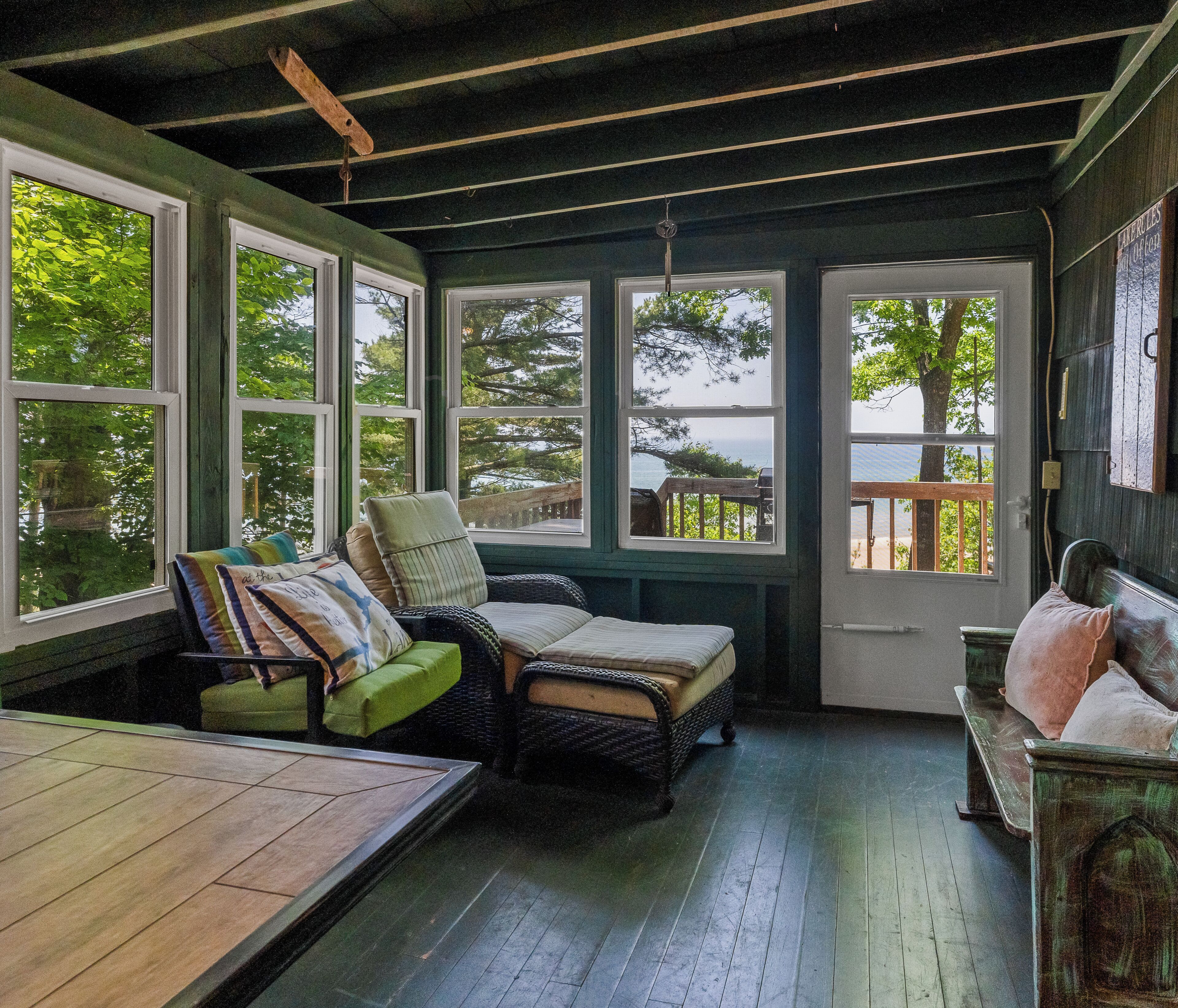 Una habitación acogedora con piso de madera, un sofá verde, una silla de mimbre y un banco. Amplias ventanas ofrecen vistas a árboles y un muelle.