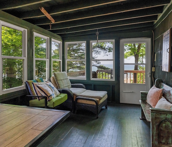 A cozy room with a wooden floor, a green sofa, a wicker chair, and a bench. Large windows offer views of trees and a dock.