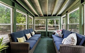 A covered porch with two blue sofas and wicker chairs, large windows, and a view of trees.