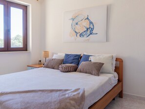 Un dormitorio con cama, almohadas, una mesita de noche, lámpara y un cuadro colgado en la pared.