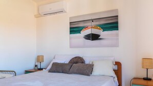Un dormitorio con cama, almohadas, mesita de noche, lámpara, un cuadro de un barco en la pared y un equipo de aire acondicionado.