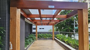 A covered outdoor walkway with wooden beams and a tiled floor, flanked by greenery and a building in the background. - Cozy Studio w Balcony & Pool in Carnival's Heart - SSA009 (Salvador - BA)