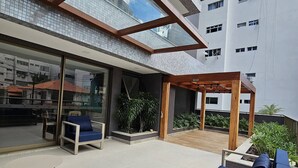 A modern outdoor area with a glass-enclosed patio, a seating area with a blue sofa, and a wooden pergola. - Cozy Studio w Balcony & Pool in Carnival's Heart - SSA009 (Salvador - BA)