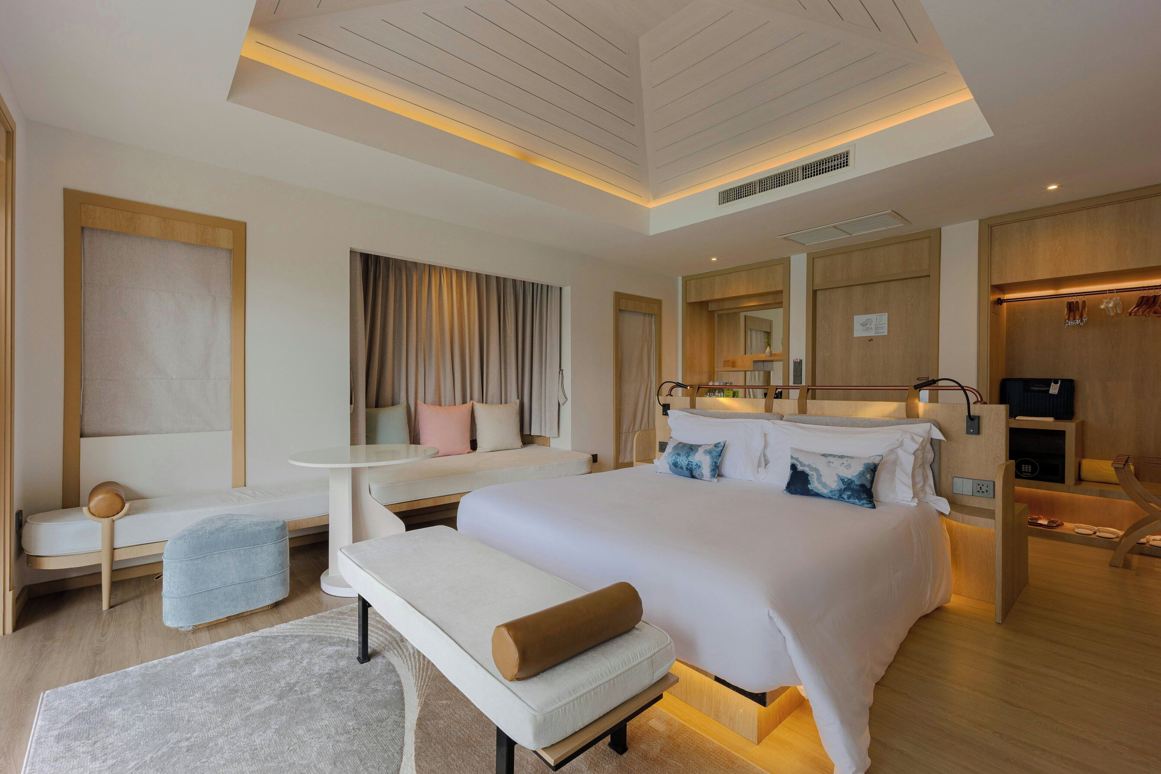 A modern hotel room with a large bed, a sitting area with a bench, a small table, and a wardrobe.