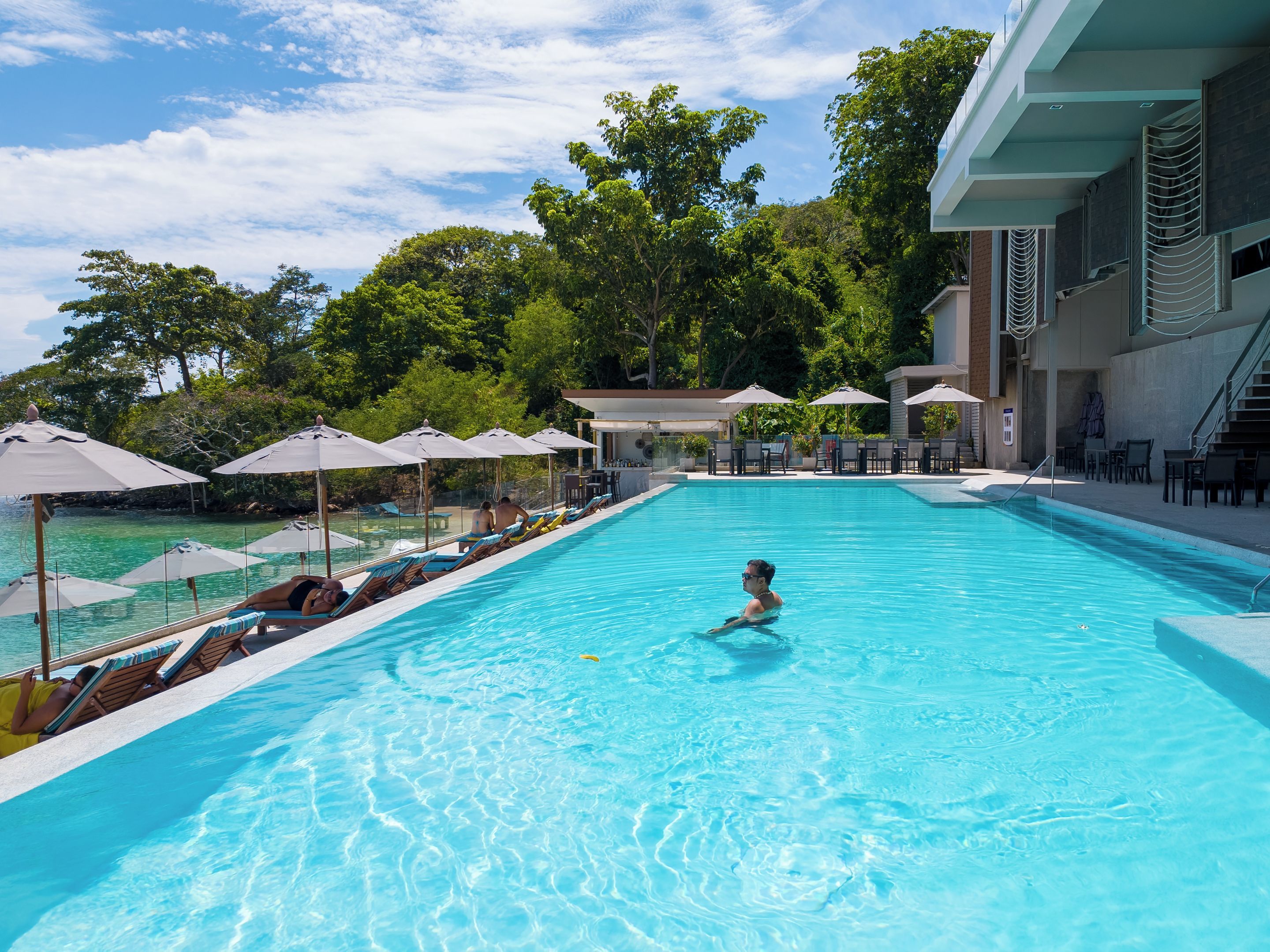 Una piscina con sillas de descanso y sombrillas, rodeada de árboles y un edificio.