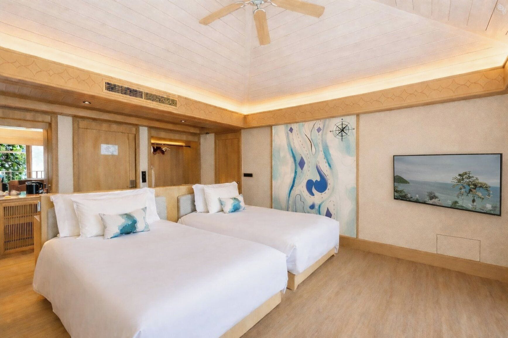 A hotel room with two beds, wooden flooring, a ceiling fan, a painting on the wall, and a view of the sea.