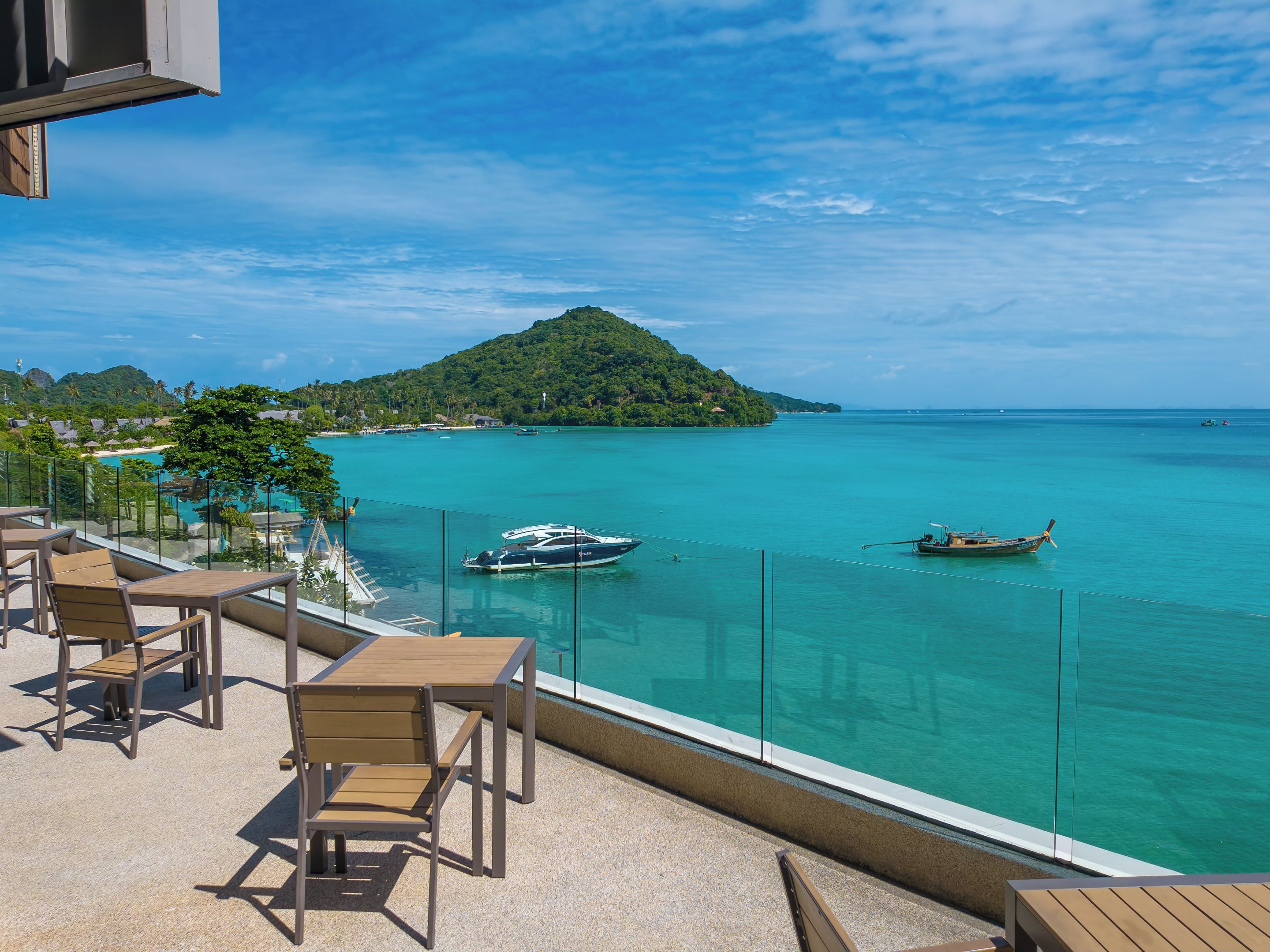 A balcony with outdoor seating overlooking a turquoise sea, a small island, and boats.