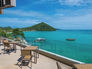 Ein Balkon mit Sitzgelegenheit mit Blick auf das türkisfarbene Meer, eine kleine Insel und Boote.