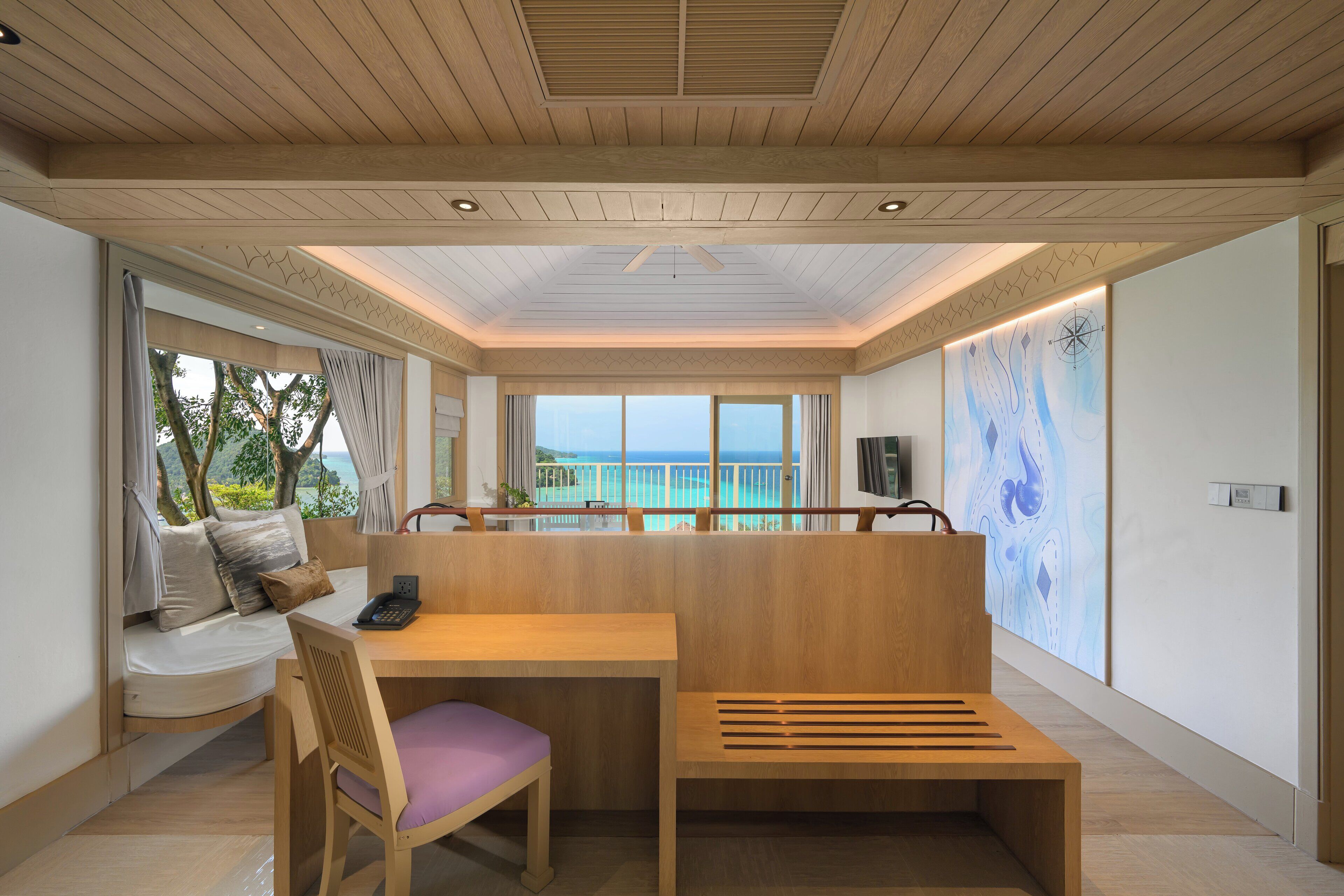 A modern room with a wooden ceiling, a view of the sea, a wooden desk, a cushioned chair, and a bench.