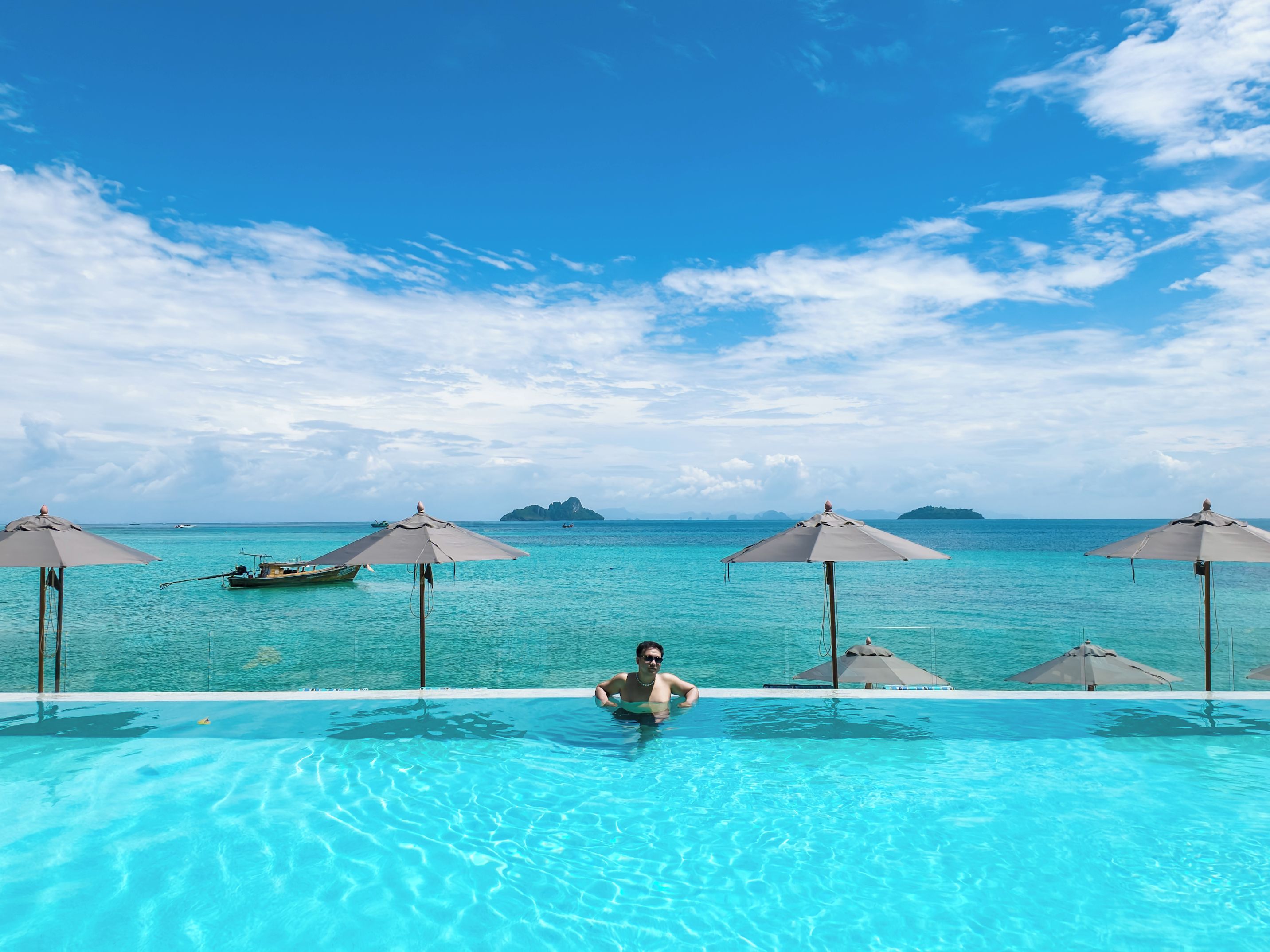 Une vue sur la piscine avec un ciel dégagé, une personne dans l’eau et plusieurs parasols.