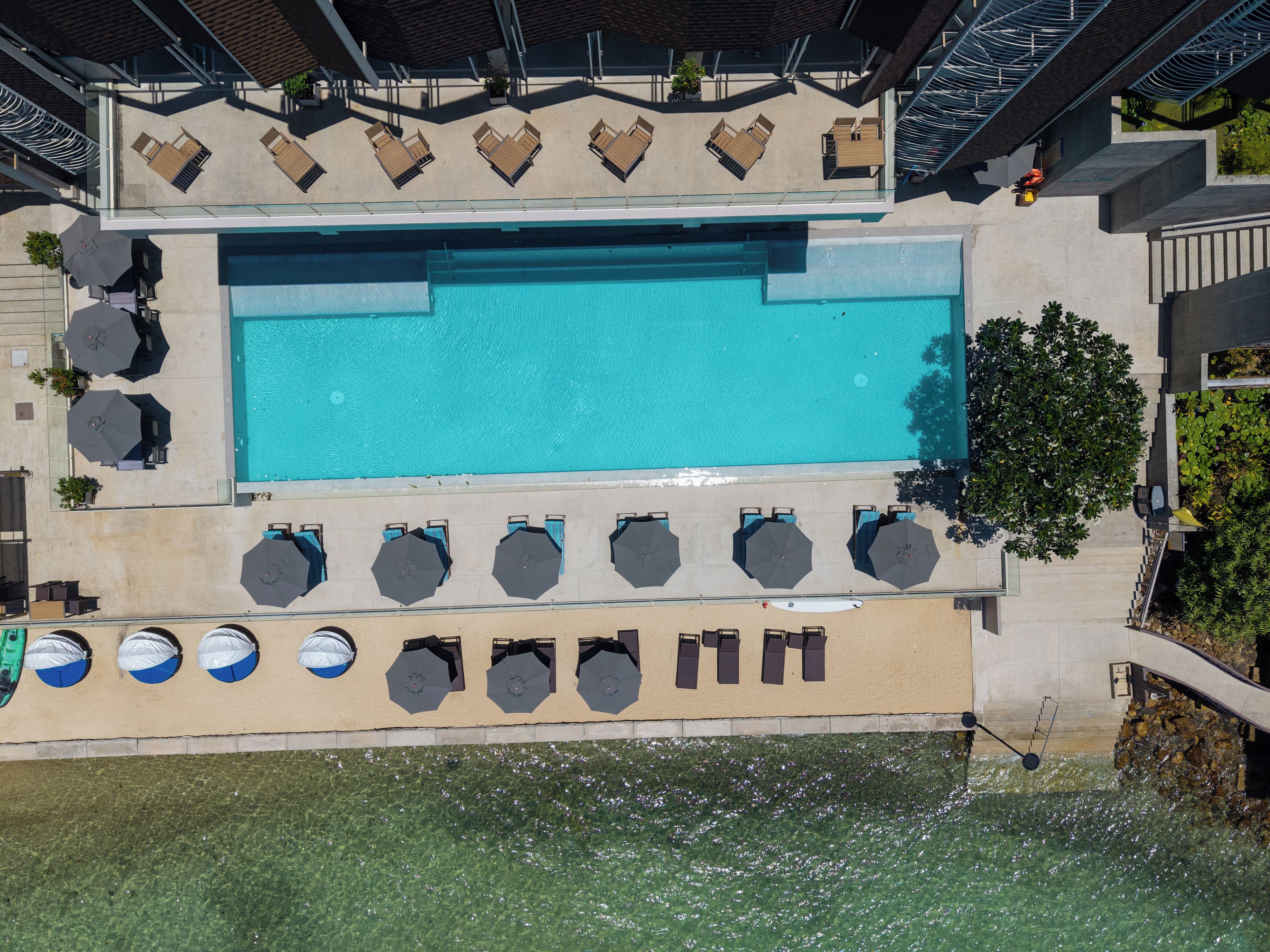 Vista aérea de uma área de piscina de hotel, com uma piscina retangular, espreguiçadeiras, guarda-sóis e uma escadaria que dá para um nível inferior.