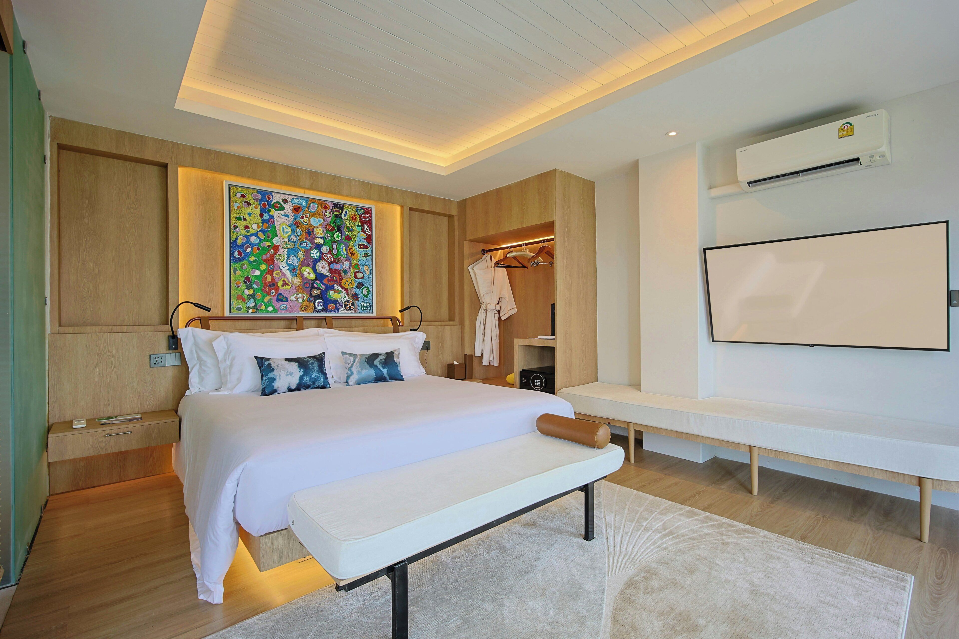 Une chambre à coucher moderne avec un grand lit, des tables de chevet, un banc, un téléviseur fixé au mur, un appareil de climatisation et une œuvre d’art colorée accrochée au mur.