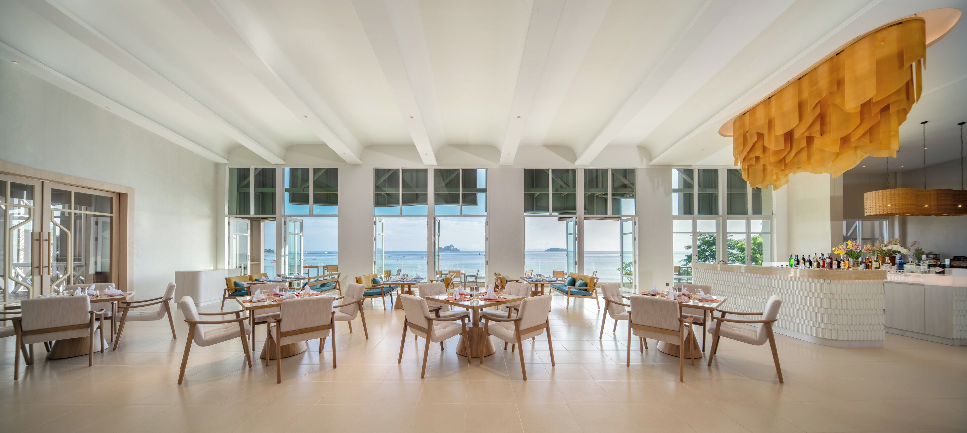 A spacious dining area with multiple tables and chairs, large windows offering ocean views, and a bar area with a unique yellow hanging light fixture.