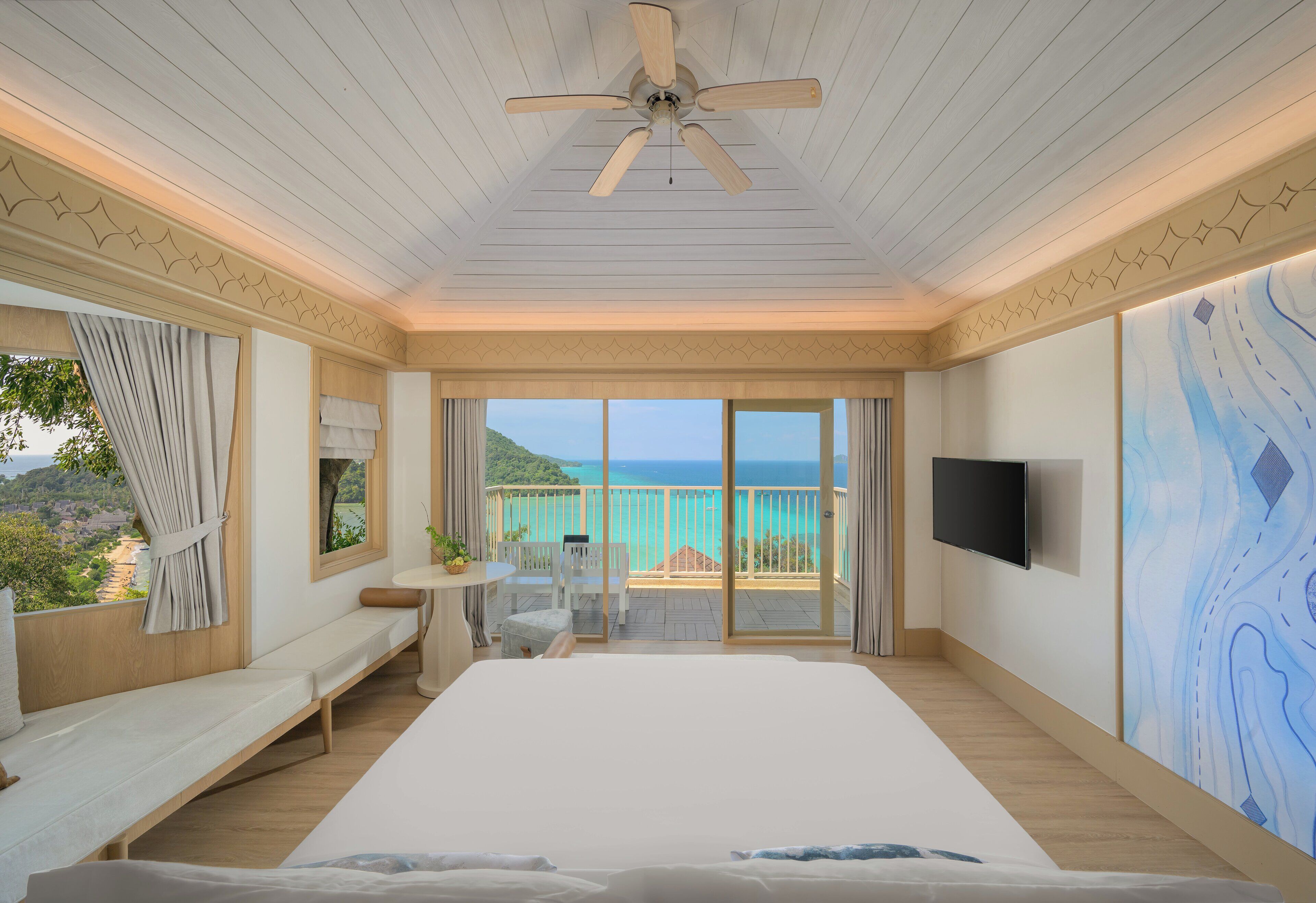 Un dormitorio amplio con una cama grande, un ventilador de techo, un balcón con vistas al mar y un televisor de pantalla plana montado en la pared.
