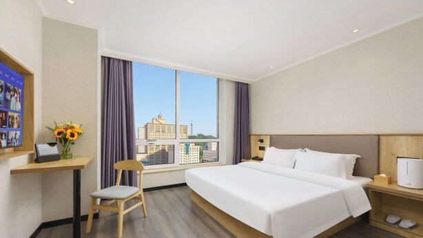 A hotel room with a large bed, a desk with a computer, a chair, a nightstand, and a window with a city view. - Dalian Youxi-ChengYuan Hotel (Dalian)
