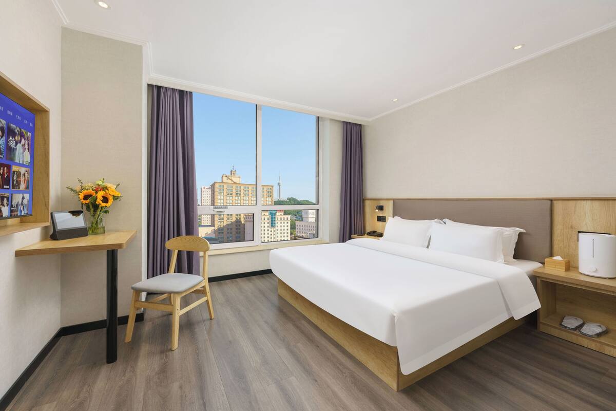 Une chambre d’hôtel avec un grand lit, un bureau avec un ordinateur, une chaise, une table de chevet et une fenêtre donnant sur la ville.