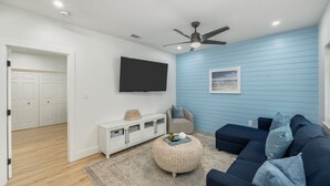 A modern living room with a blue wall, a ceiling fan, a flat-screen TV, a dark blue sofa, and a white entertainment unit. - With Hot Tub & Mini Golf | Sunny Haven #617 (Fort Walton Beach)