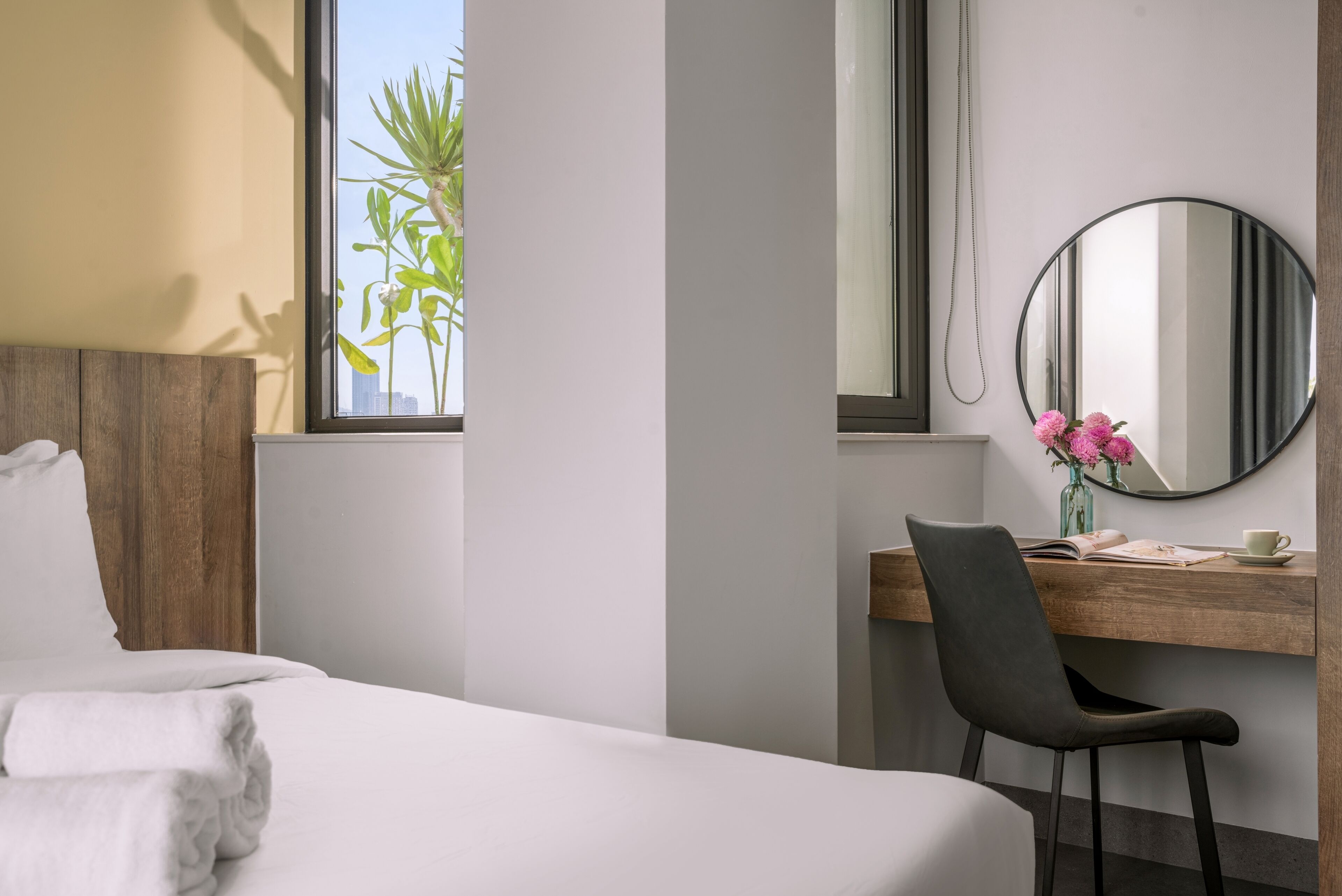 A hotel room with a bed, a desk with a chair, a round mirror, and a window with a view of greenery.