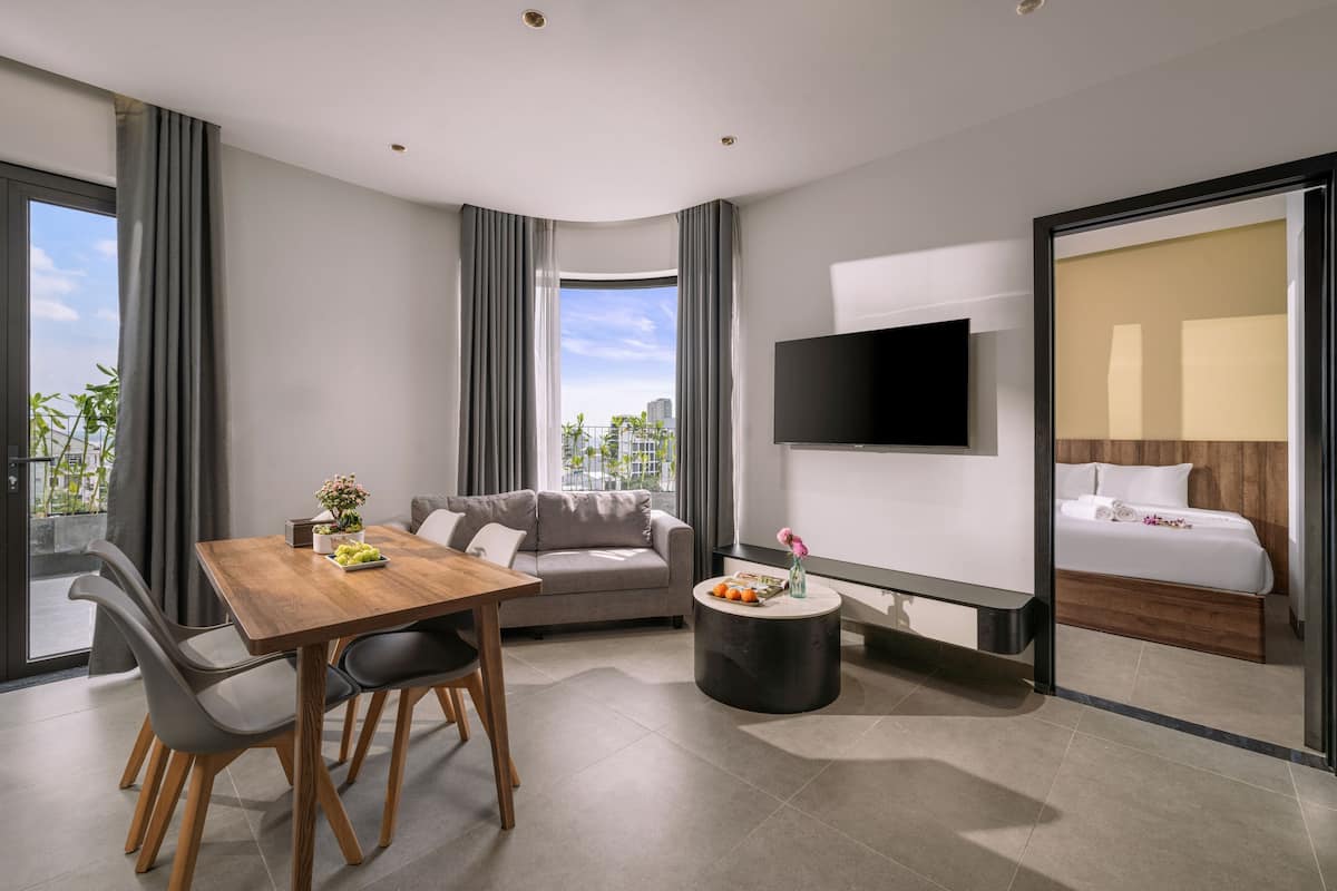 A modern hotel room with a dining area, a sofa, a round coffee table, a bed, and a TV.
