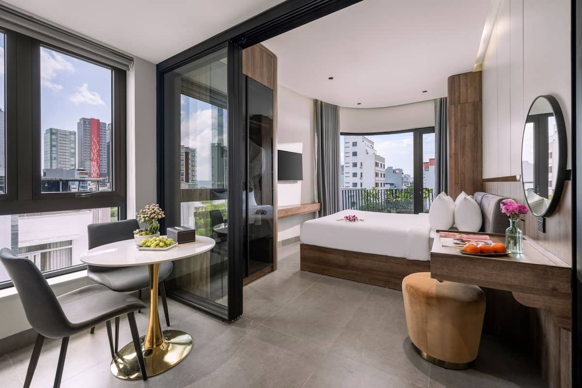 A modern hotel room with a large window, a dining area, a bed, and a small bathroom.