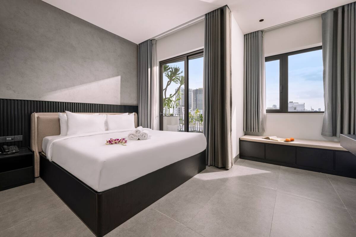 A modern hotel room with a large bed, a bedside table, a window with curtains, and a view of the cityscape.