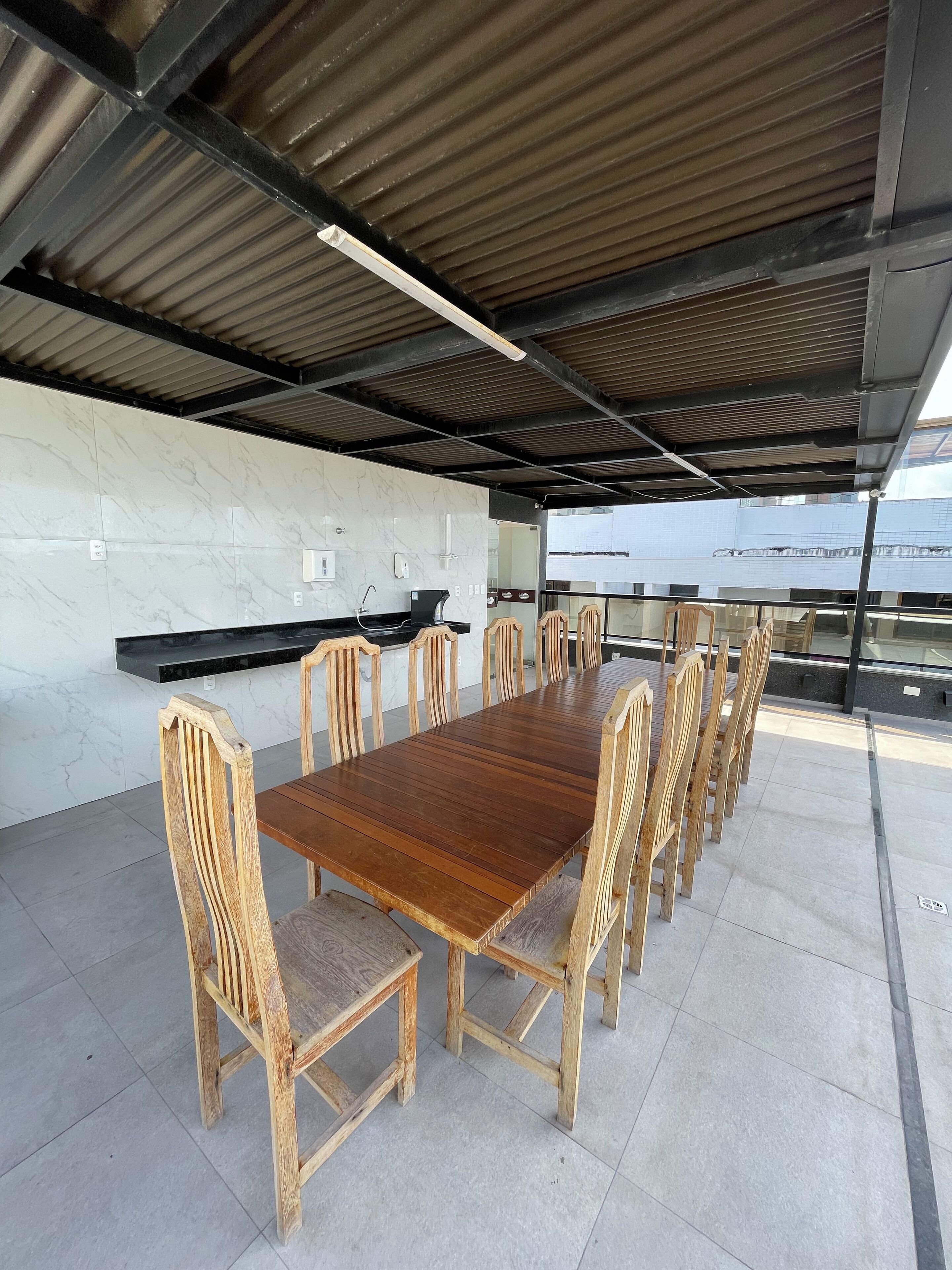 Een overdekt terras met een lange houten tafel en bijpassende stoelen, een ingebouwde barbecue en een witte betegelde muur.