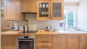 A modern kitchen with wooden cabinets, a Bosch oven, a kettle, and a window with a view of trees. - Castle Cottage, Orford (Orford)