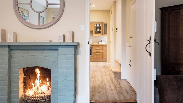 A room with a fireplace, a mirror, and a view into a kitchen area. - Castle Cottage, Orford (Orford)