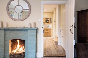 A room with a fireplace, a mirror, and a view into a kitchen area. - Castle Cottage, Orford (Orford)