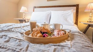 A neatly made bed with a tray of breakfast items including croissants, a glass of milk, and pastries, set in a room with a wooden headboard and two bedside lamps. - Castle Cottage, Orford (Orford)