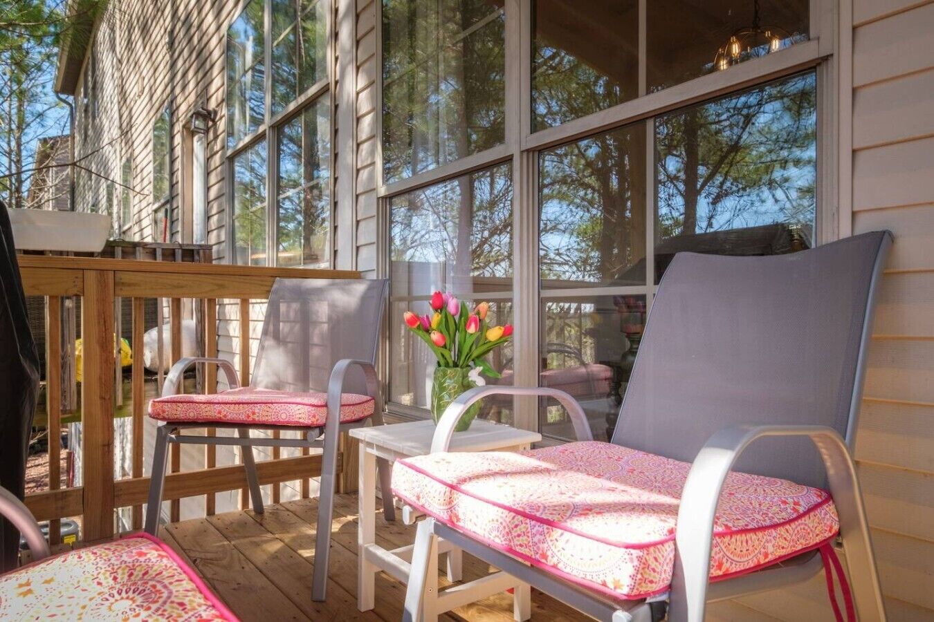 Um alpendre coberto com duas cadeiras, uma mesa, um vaso com flores e vista para árvores através de grandes janelas.