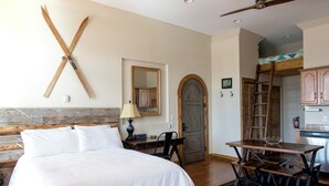 A bedroom with a wooden headboard, a bed with white linens, a desk with a lamp, and a dining table with benches. - Studio 508 Downtown Leadville (Leadville)