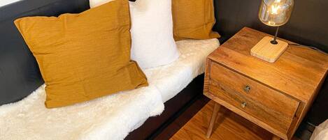 A corner bed with a white fluffy bedspread, two mustard-colored pillows, and a wooden nightstand with a lamp.
