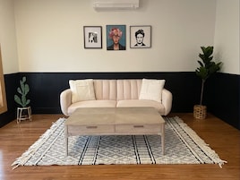 A living room with a beige sofa, a coffee table, and a patterned rug. There are three framed artworks on the wall.