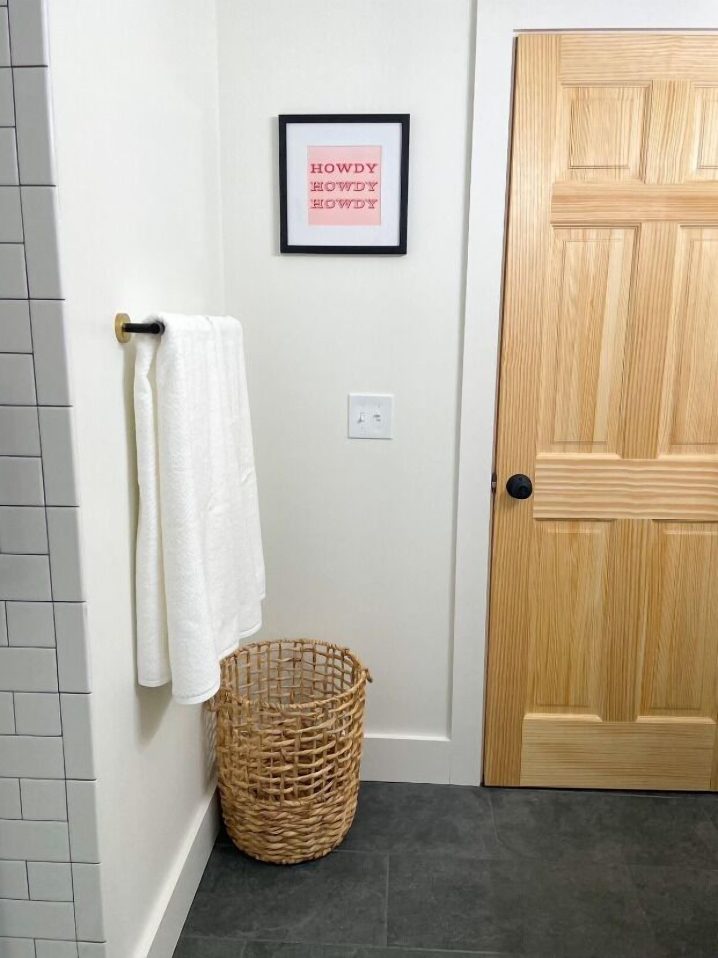A bathroom with a white tiled wall, a wooden door, a framed picture with text, a towel rack with a white towel, and a wicker basket.