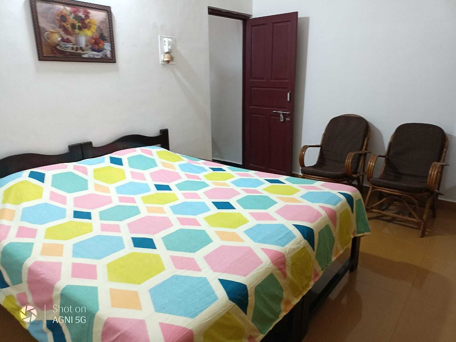 A room with a bed, two chairs, a door, and a wall-mounted light switch.