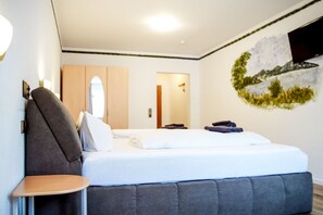 A hotel room with a bed, a chair, a wall-mounted TV, and a door leading to another room. - Comfortable and versatile room -perfect for relaxation, work and exchange (Winterberg)