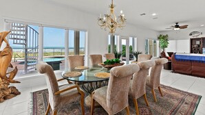 En rummelig stue med et glasbord omgivet af beige polstrede stole, en lysekrone og havudsigt gennem store vinduer.