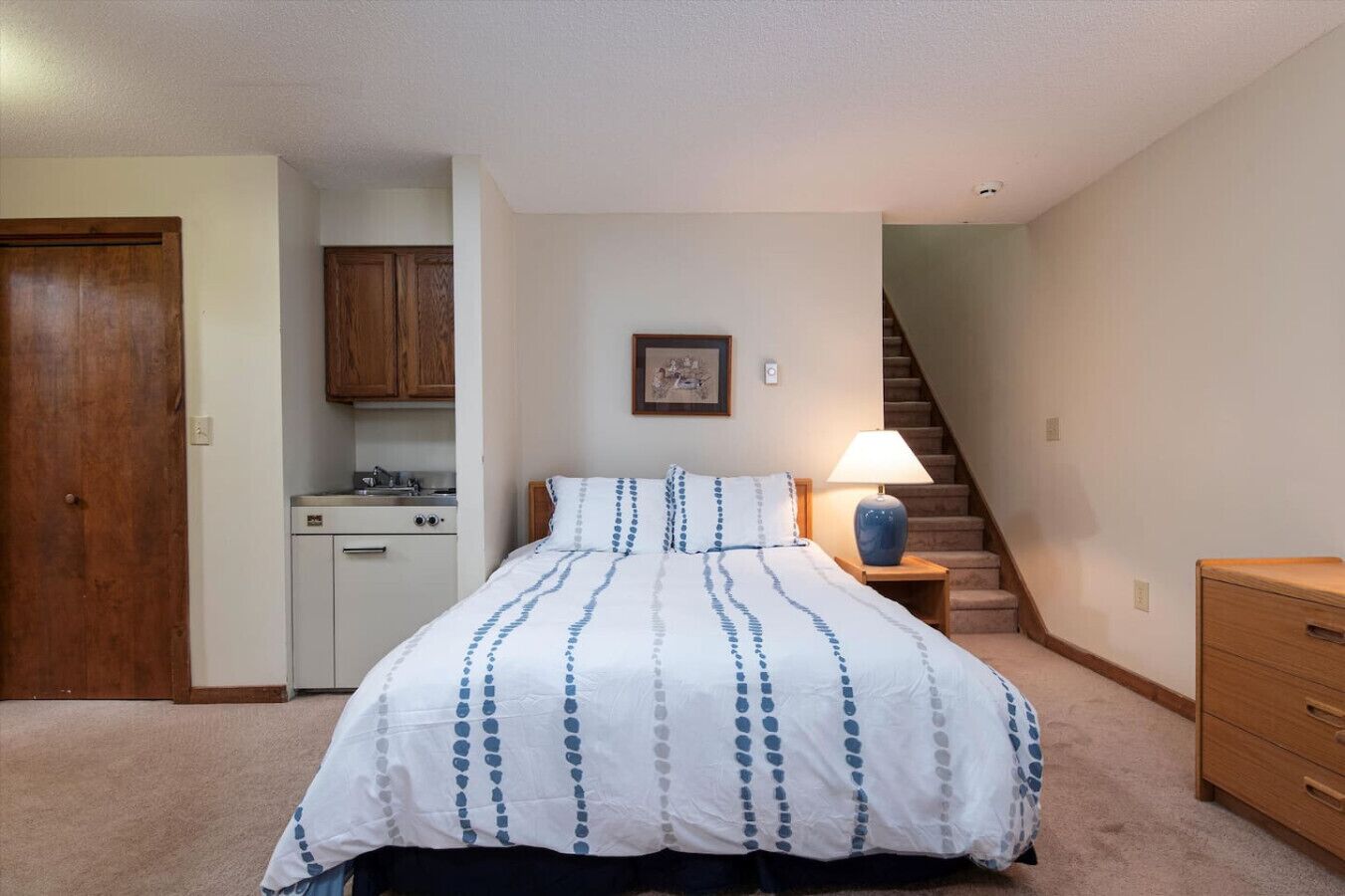 A bedroom with a bed, a nightstand with a lamp, a wooden dresser, a kitchen area with cabinets, and a staircase.
