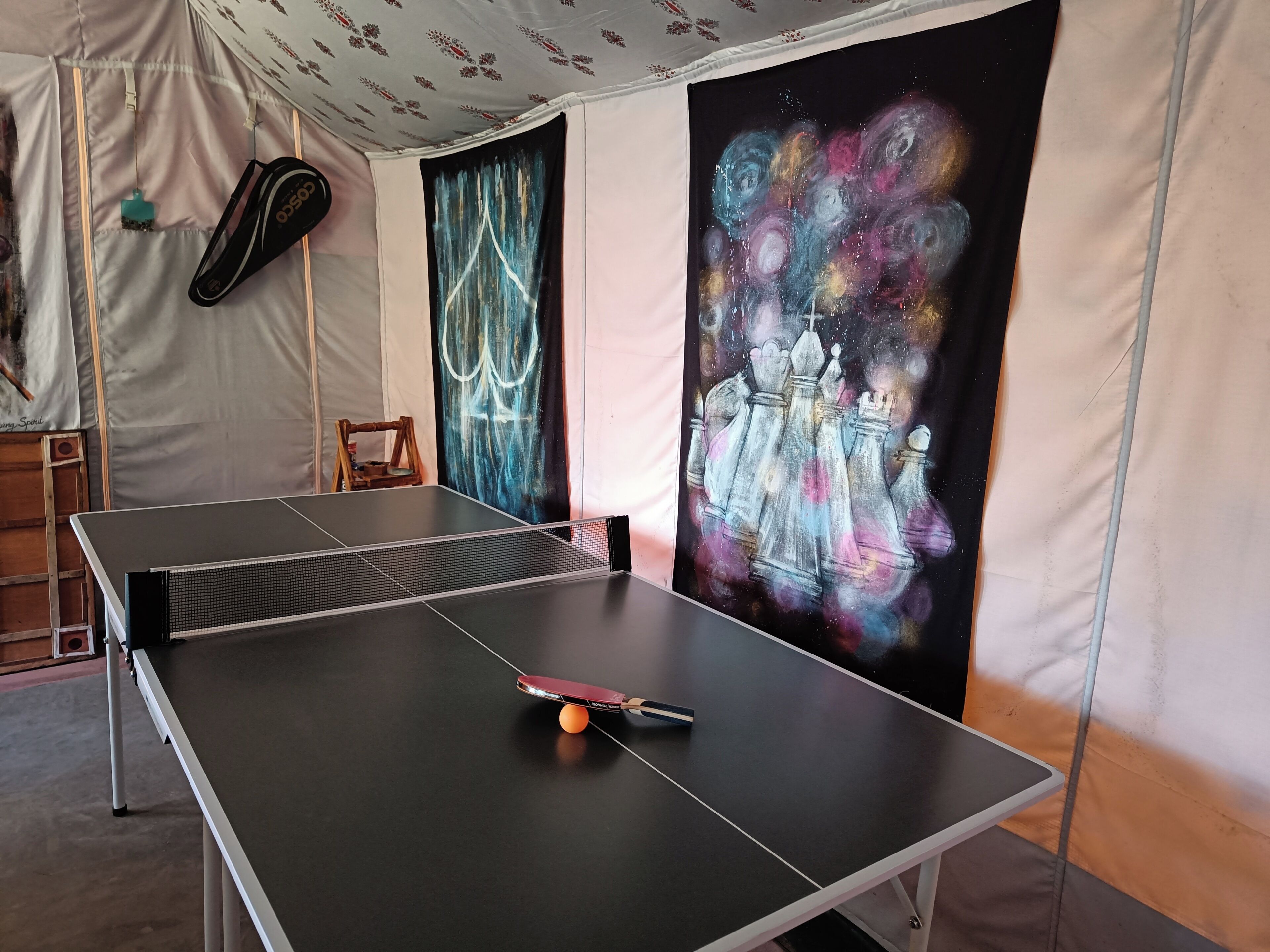 Una mesa de ping pong con una paleta de tenis de mesa, una silla de madera y una pared con un cuadro con temática de galaxia.