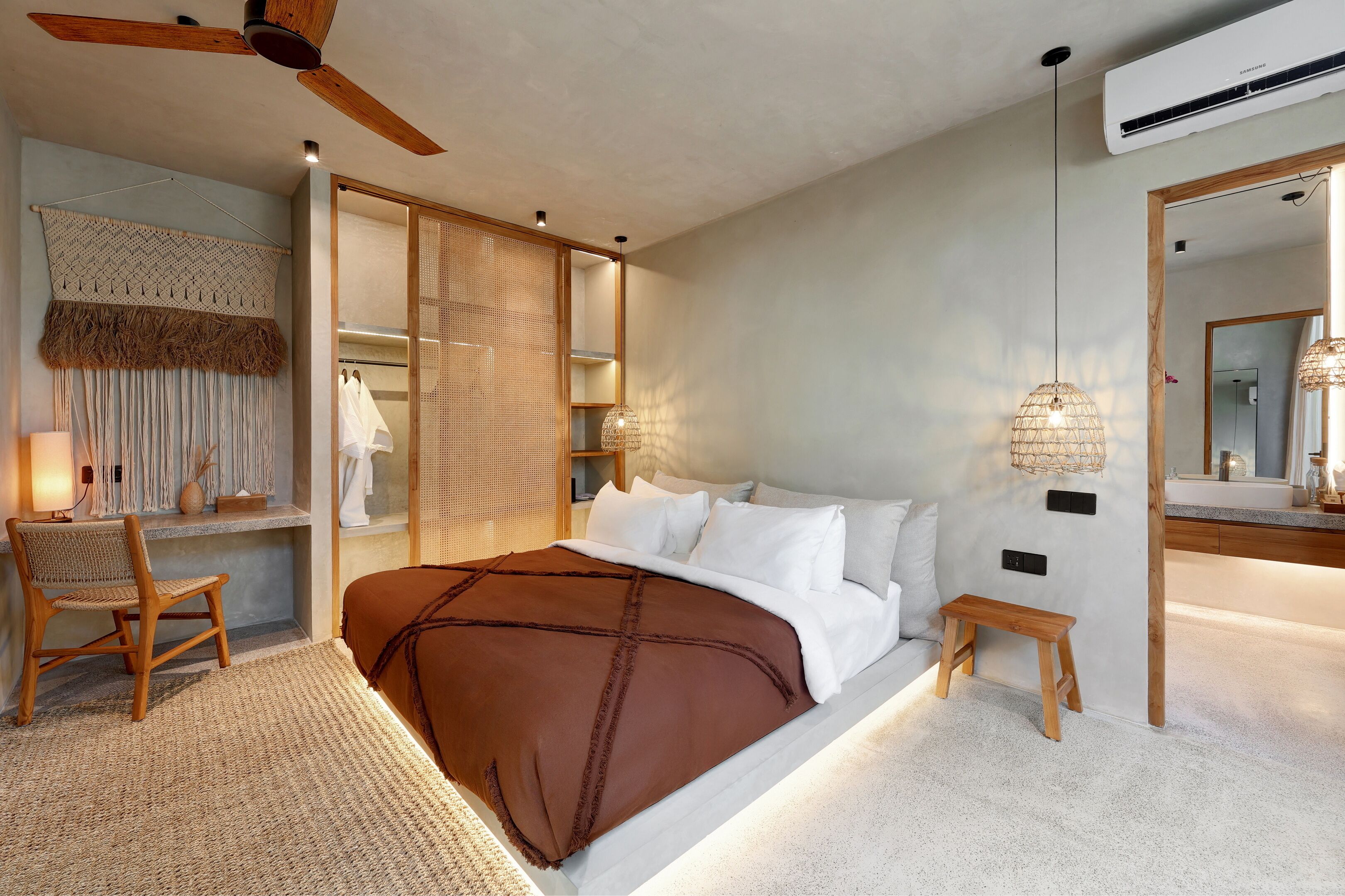Un dormitorio moderno con una cama grande, una silla de madera, un taburete pequeño, un ventilador de techo y un aire acondicionado instalado en la pared.