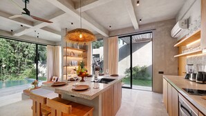 A modern kitchen with a central island, wooden chairs, a hanging light fixture, and a view of a swimming pool. - Garden Villa Ubud By BREIG (Ubud)