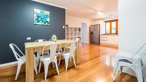 A dining area with a wooden table and white chairs, a kitchen in the background, and a dark wall with a blue abstract painting.