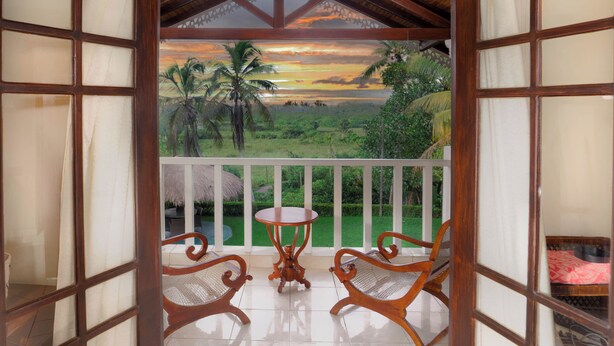 Un balcon en bois avec une table et deux chaises, donnant sur un paysage luxuriant parsemé de palmiers et illuminé par un coucher de soleil.