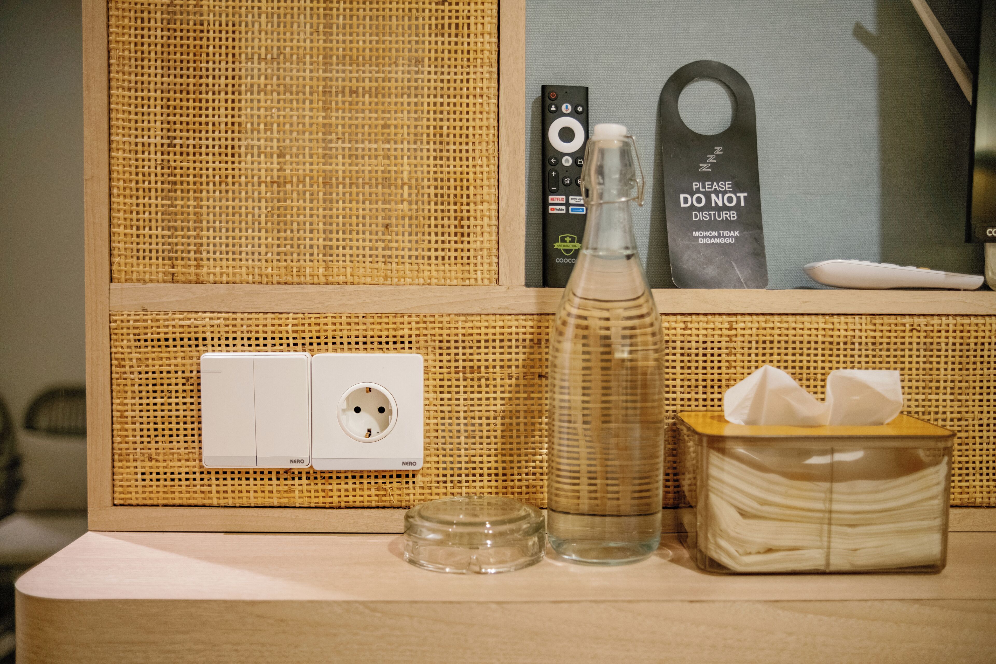 A hotel room with a woven wall panel, a power outlet, a remote control, a glass bottle, and a tissue box.