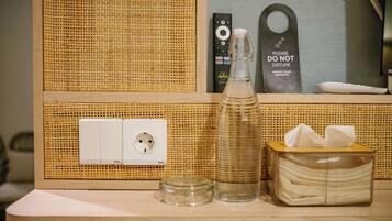 A hotel room with a woven wall panel, a power outlet, a remote control, a glass bottle, and a tissue box.