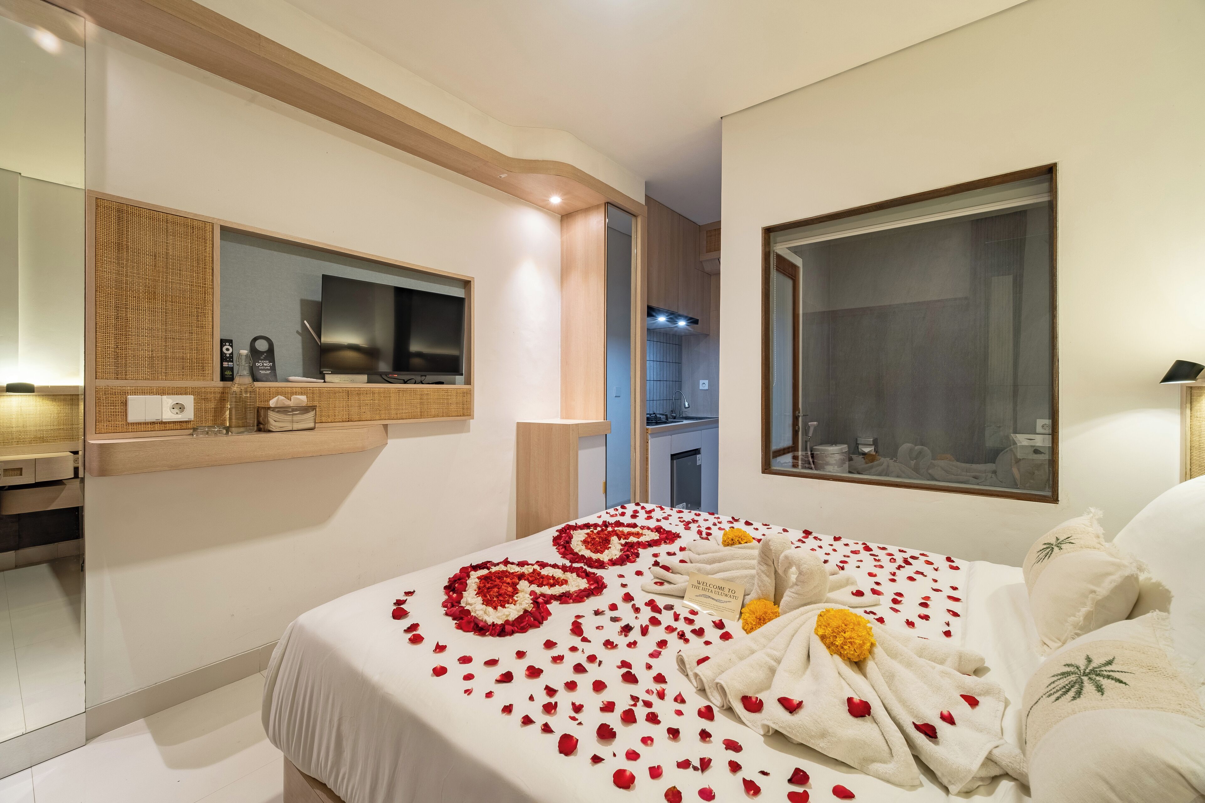 A hotel room with a bed adorned with rose petals and a welcome mat, a large mirror, and a view of the kitchen.