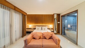 Une chambre d’hôtel moderne dotée d’un grand lit, d’un canapé, d’une salle de bain fermée par des parois de verre et de lambris en bois.