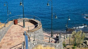 Uma plataforma circular com pavimento de pedra, guarda-corpos de metal e um muro de pedra, com vista para o mar azul.